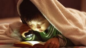young child reading a book under a blanket with a flashlight