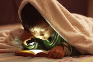 young child reading a book under a blanket with a flashlight